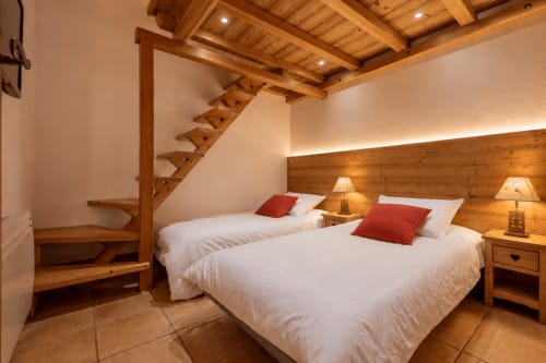 Chambre alpine cosy avec poutres en bois, deux lits aux oreillers rouges, tables de nuit en bois et plafond apparent à Cosyarc Arc 1800