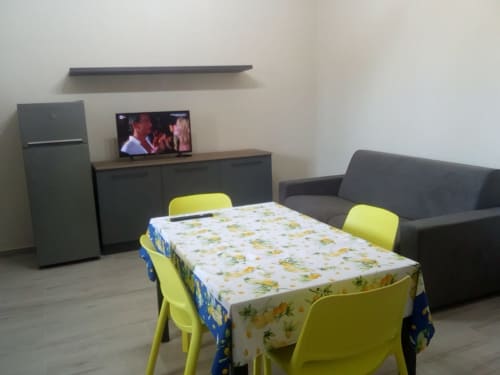 Zona living dell'appartamento Amelia Blue a My Home Apulia con sedie gialle, tavolo, divano, TV, angolo cottura e frigorifero