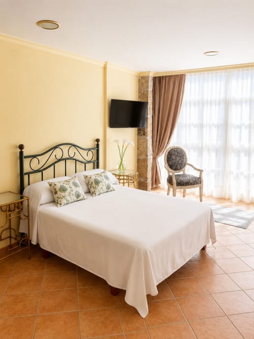 Luminoso dormitorio de estudio en Baiona con cama de hierro forjado, ropa de cama blanca, paredes amarillas, TV y balcón amueblado