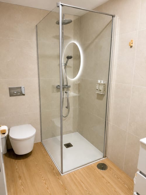 Banheiro moderno com chuveiro, cascata de água, espelho iluminado, vaso sanitário branco e piso de madeira em Terra