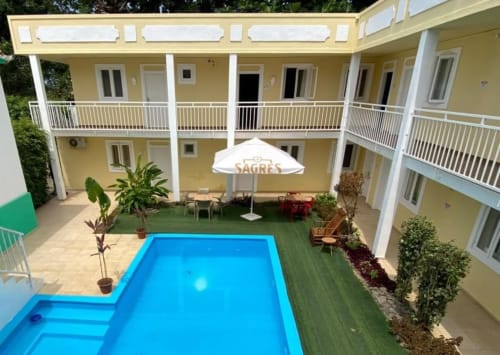 Pátio do Hotel PZero em Albufeira com piscina azul, edifício amarelo, corrimãos brancos, guarda-sol e gramado verde