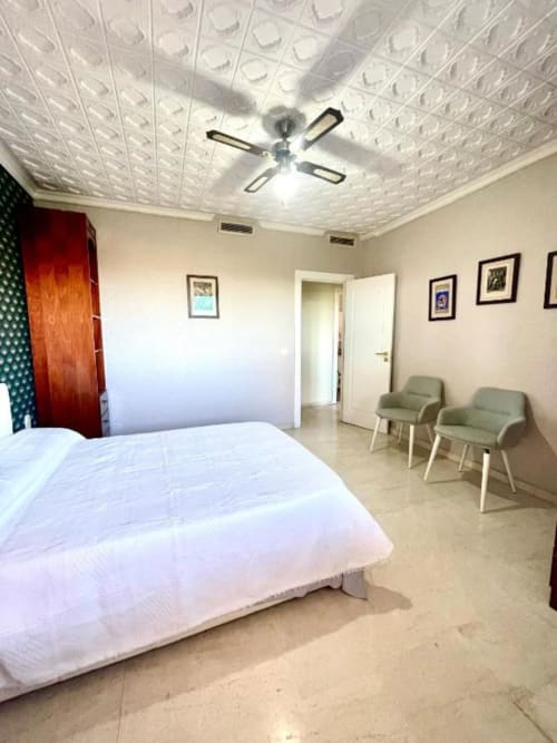 Dormitorio moderno con cama blanca, ventilador de techo, sillas verdes y cuadros en Villa Magos, Málaga