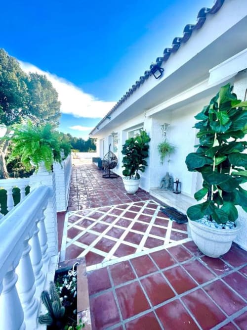 Soleada terraza de Villa Magos en Málaga con edificio blanco, suelo de baldosas rojas, plantas en macetas y vistas al océano