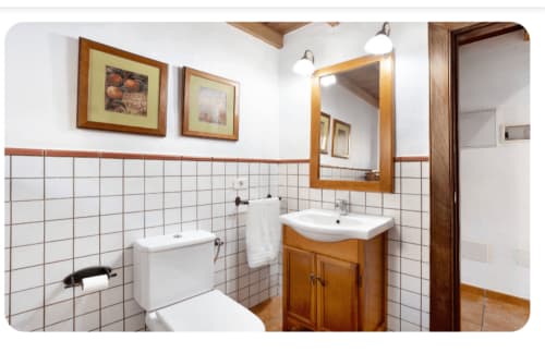 Luminoso cuarto de baño en la villa Casa Rural La Caldera con paredes de azulejos blancos, mueble de madera, espejo, inodoro y suelo de barro