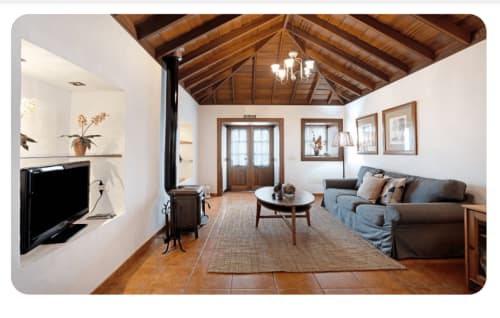 Amplia sala de estar con vigas de madera vista, chimenea, sofá gris y cuadros en villa de La Palma