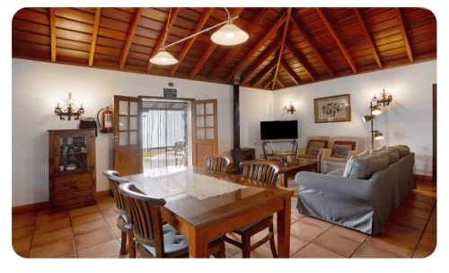 Sala de Casa Rural Los Llanos Negros con techo de madera, mesa de comedor, TV, sofá azul, suelo de barro, vistas al mar