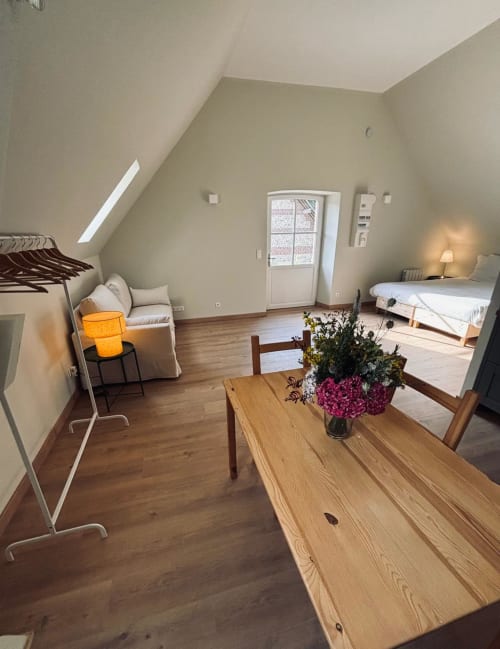 Grand Studio du Manoir de Daubeuf : chambre mansardée lumineuse avec table en bois, lit, coin salon et fleurs