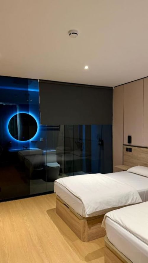 Habitación doble moderna en Hotel ITOUR con dos camas, espejo circular iluminado en azul, baño con cristalería y suelo de madera