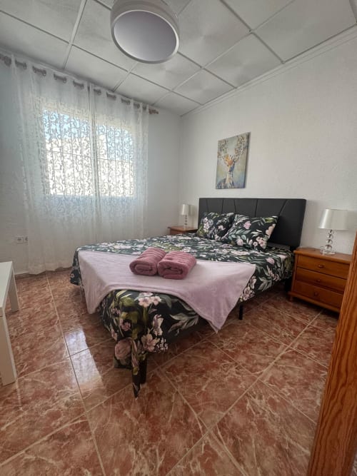 Dormitorio amplio en la casa de vacaciones Haus Holiday Homes en Los Alcázares con ropa de cama floral, mesita de noche de madera y suelo de barro cocido