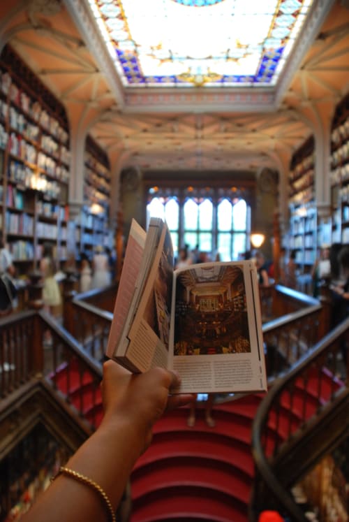 Mão segurando guia turístico em biblioteca ornada de Aveiro com vitral na claraboia e tapete vermelho