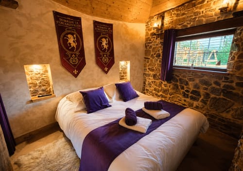 Chambre confortable à La Synchronicité B&B à Saint Sauveur : murs en pierre, lit blanc, touches violettes et bannières héraldiques