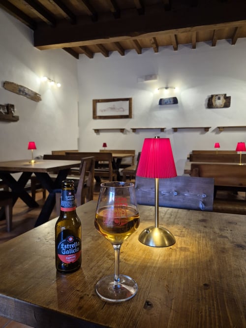 Barra rústica de Aldea Rural Pazos De Arenteiro con cerveza Estrella Galicia, copa de vino y lámpara rosa sobre mesa de madera