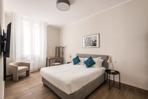 Camera moderna dell'hotel Flavia a Roma con letto grigio, biancheria bianca, cuscini turchesi, pavimenti in legno, TV e poltrona beige