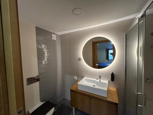 Banheiro moderno da RISOTURISMO com espelho redondo iluminado, lavatório branco, paredes de azulejo cinza e gabinete de madeira