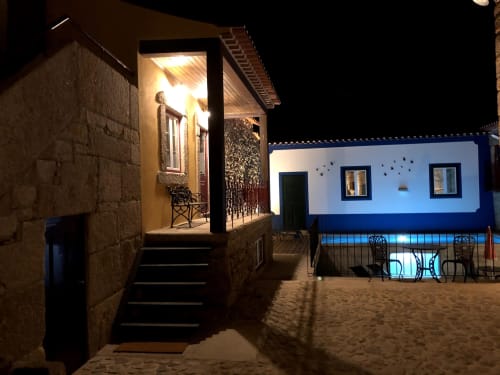 Vista noturna da entrada da quinta RISOTURISMO com edifício de pedra iluminado e casa pintada de azul em solo arenoso