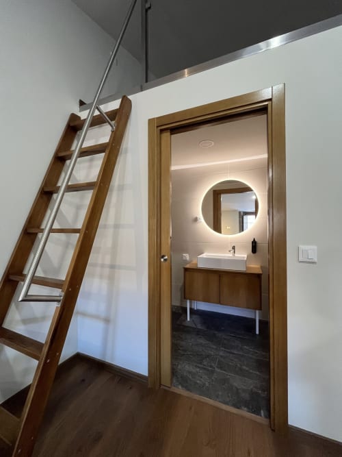 Entrada do banheiro moderno com porta de madeira, espelho redondo iluminado, cuba de apoio e escada tipo sótão no Quarto Massena em