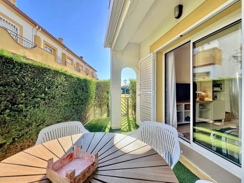 Terraza soleada de la casa Estartit con sillas blancas, tarima de madera, seto frondoso y puertas de vidrio abiertas