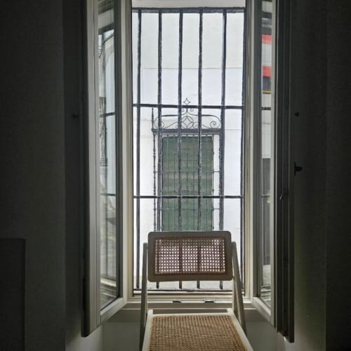 Puertas abiertas al balcón de un apartamento en Vejer con barandillas de hierro forjado, persianas verdes y silla tejida con vistas a la ciudad.
