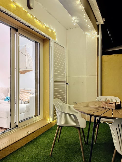 Patio de cabaña moderna en Estartit con mesa de comedor, sillas blancas, césped artificial y luces de cuerda brillantes