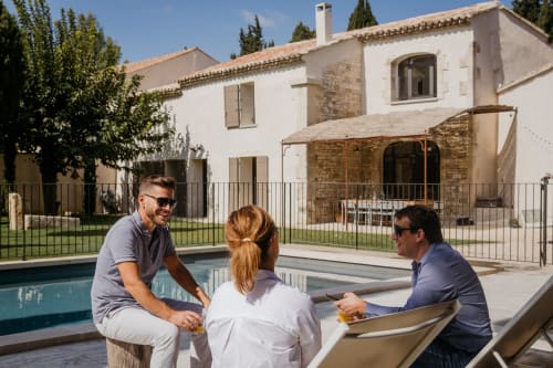 Trois clients savourent leurs boissons au bord de la piscine du Mas des Ecoliers, une maison d'hôtes provençale à Châteaurenard aux murs de pierre rustique.