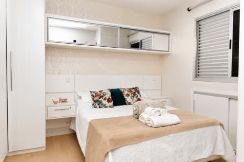Quarto moderno do apartamento CSS em Matinhos com cama queen, almofadas florais, espelho e janela com venezianas brancas