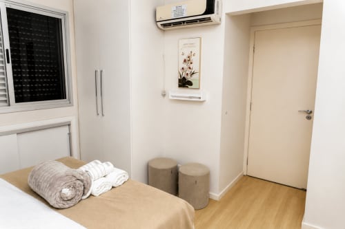 Quarto moderno da CSS BnB em Matinhos com cama, roupas de cama enroladas, dois pufes, ar-condicionado e arte botânica na parede