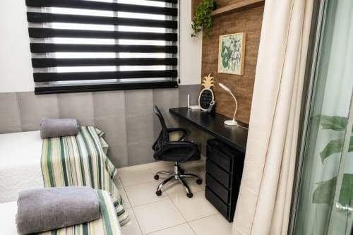 Quarto moderno da CSS BnB Matinhos com mesa de trabalho, cadeira preta, roupa de cama listrada e persianas