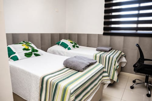 Aconchegante quarto com duas camas na CSS Matinhos BnB, cabeceira cinza, roupa de cama branca, almofadas verdes, manta listrada e cadeira de escritório
