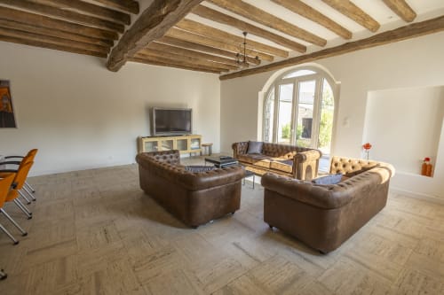 Grand salon de la Villa Chateaudine avec canapés Chesterfield en cuir, poutres apparentes, téléviseur et portes-fenêtres cintrées sur jardin