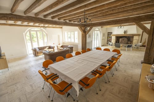 Salle à manger de la Villa Chateaudine avec table longue, chaises orange, poutres en bois, cheminée et vue sur le jardin