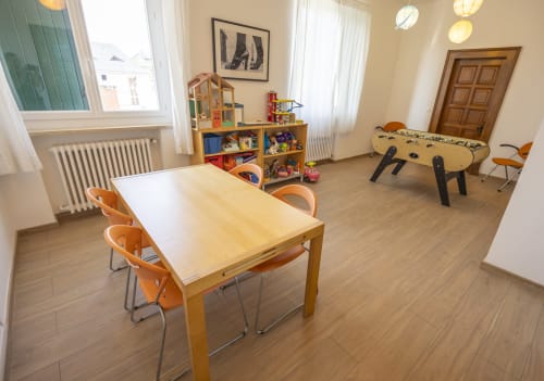 Salle de jeux lumineuse à la Villa Chateaudine avec table en bois, chaises orange, baby-foot et étagères de rangement pour jouets.
