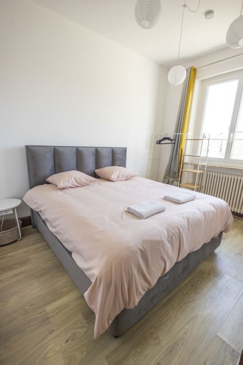 Chambre moderne de la villa Accolade à Châteaudun : lit capitonné gris, linge de lit rose et vue sur le jardin