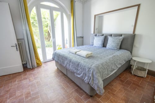 Chambre moderne de la Villa Chateaudine avec portes-fenêtres sur le jardin, lit gris, miroir et carrelage en terre cuite