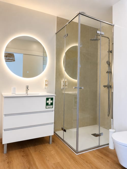 Banheiro moderno com espelho circular iluminado, pia branca, box de vidro e piso em madeira no Terra Clara