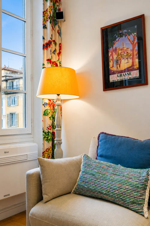 Cozy apartment corner at Enjoy Nice hotel in Nice with upholstered chair, yellow lamp, and vintage Grasse travel poster