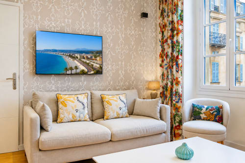 Cozy apartment living room at Enjoy Nice with beige sofa, colorful pillows, and wall-mounted TV displaying Nice coastal view