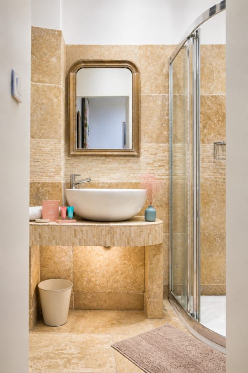 Modern bathroom in Enjoy Nice's Rossetti apartment with beige travertine tile, vessel sink, gold-framed mirror, and curved