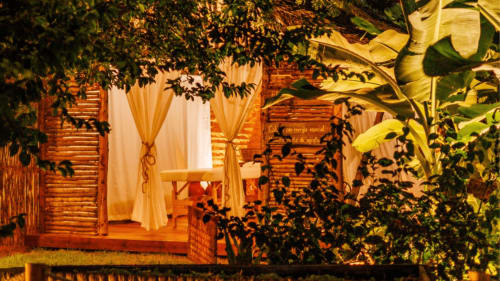 Cabana rústica na La Cozinha Pousada com iluminação aconchegante, cortinas brancas e vegetação frondosa em Cajueiro da Praia