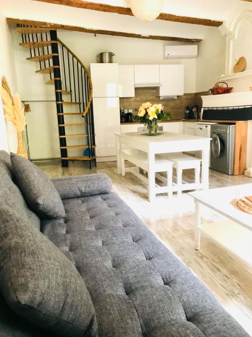 Área de estar abierta con sofá gris, mesa blanca, cocina y escalera de caracol en la suite del cottage de Torreilles