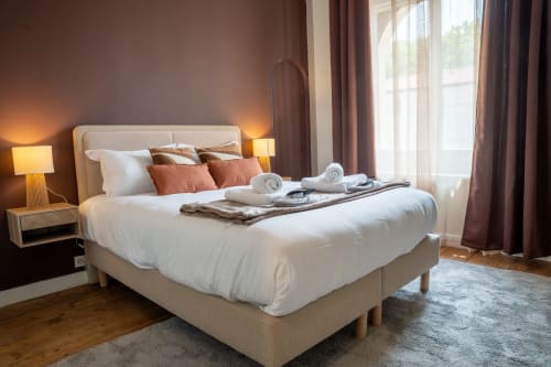 Chambre élégante au 1 Péraudel avec lit crème, oreillers terre cuite, parquet et vue sur la ville par grandes fenêtres