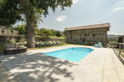 Área de piscina da Quinta de Carvalhal com espreguiçadeiras, casarão de pedra, árvore frondosa e vista para a zona rural de Guimarães