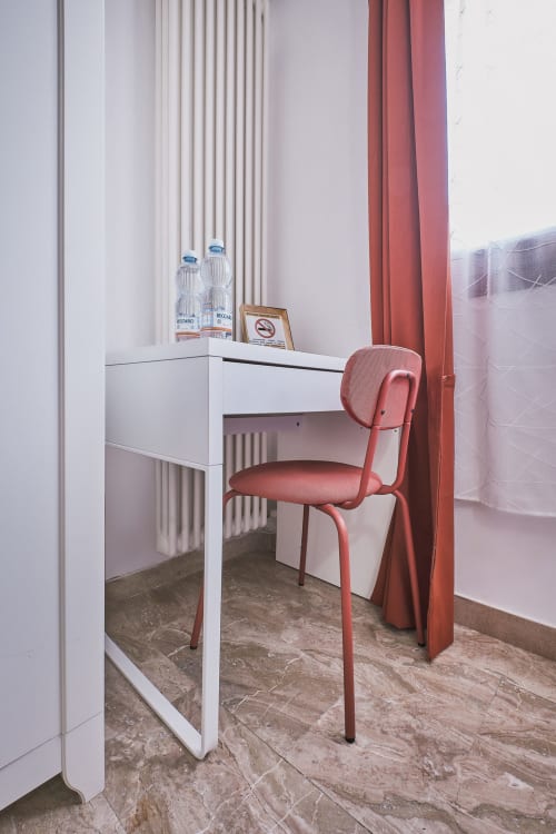 Spazio di lavoro moderno a Casa Mia B&B con scrivania bianca, sedia corallo e vista sulla città attraverso tende rosse