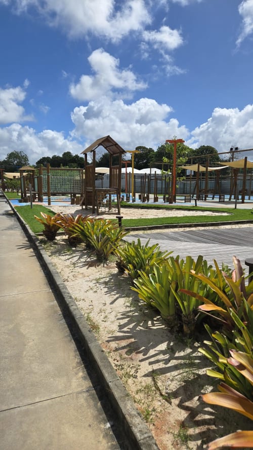 Playground moderno com equipamentos de madeira em Labedi Apartamentos em Maceió, plantas tropicais e céu azul