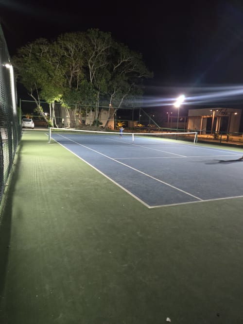 Quadra de tênis iluminada no Labedi Apartamentos em Maceió com rede acesa e árvores ao redor à noite