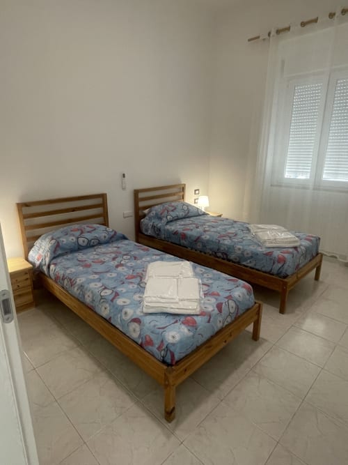 Camera da letto luminosa con due letti gemelli in legno con biancheria a fantasia, tende bianche e pavimento in piastrelle a Termoli,