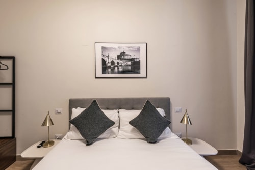 Camera da letto moderna presso la casa vacanze di Flavia a Roma con testata grigia, biancheria bianca, cuscini scuri, lampade in ottone e cornici