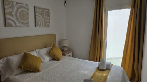 Dormitorio moderno con acentos dorados, cabecera beige, ropa de cama blanca, almohadas amarillas y vistas al jardín desde las puertas de la terraza