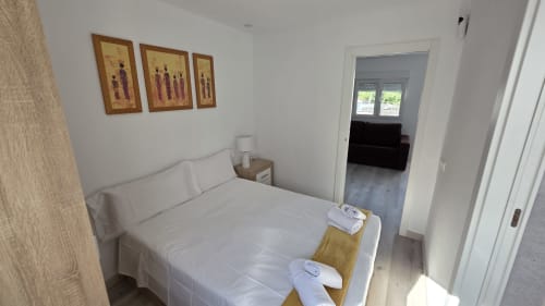 Dormitorio luminoso en bungalow frente al mar en Oropesa con cama blanca, mesita de noche de madera, cuadros enmarcados y vista al jardín