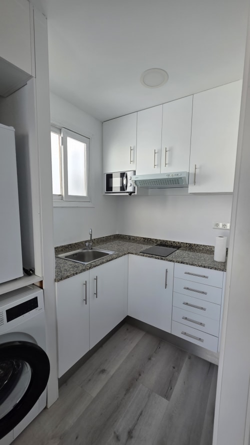 Cocina compacta moderna en apartamento de Benicàssim con gabinetes blancos, encimera de granito, microondas y fregadero de acero inoxidable