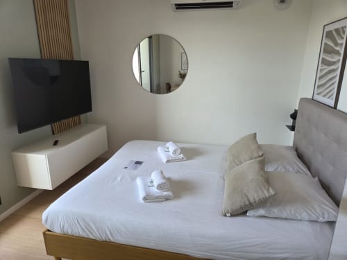 Chambre d'hôtel moderne aux Loges de la Mer, Pérols avec lit blanc, TV écran plat, miroir rond et décor minimaliste